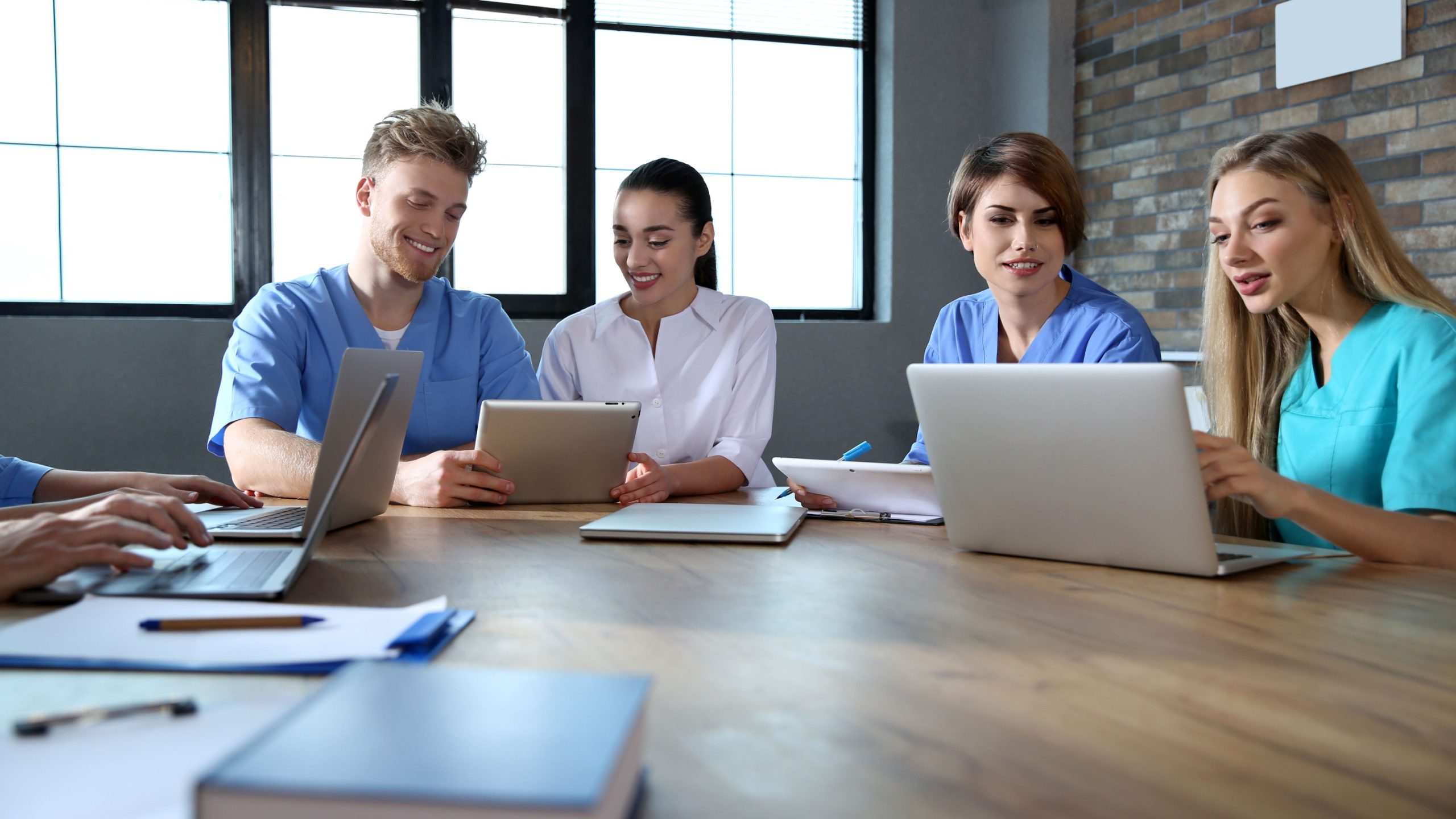 A group of medical school students wearing scrubs, sitting at a large desk with laptops and study books in group study.